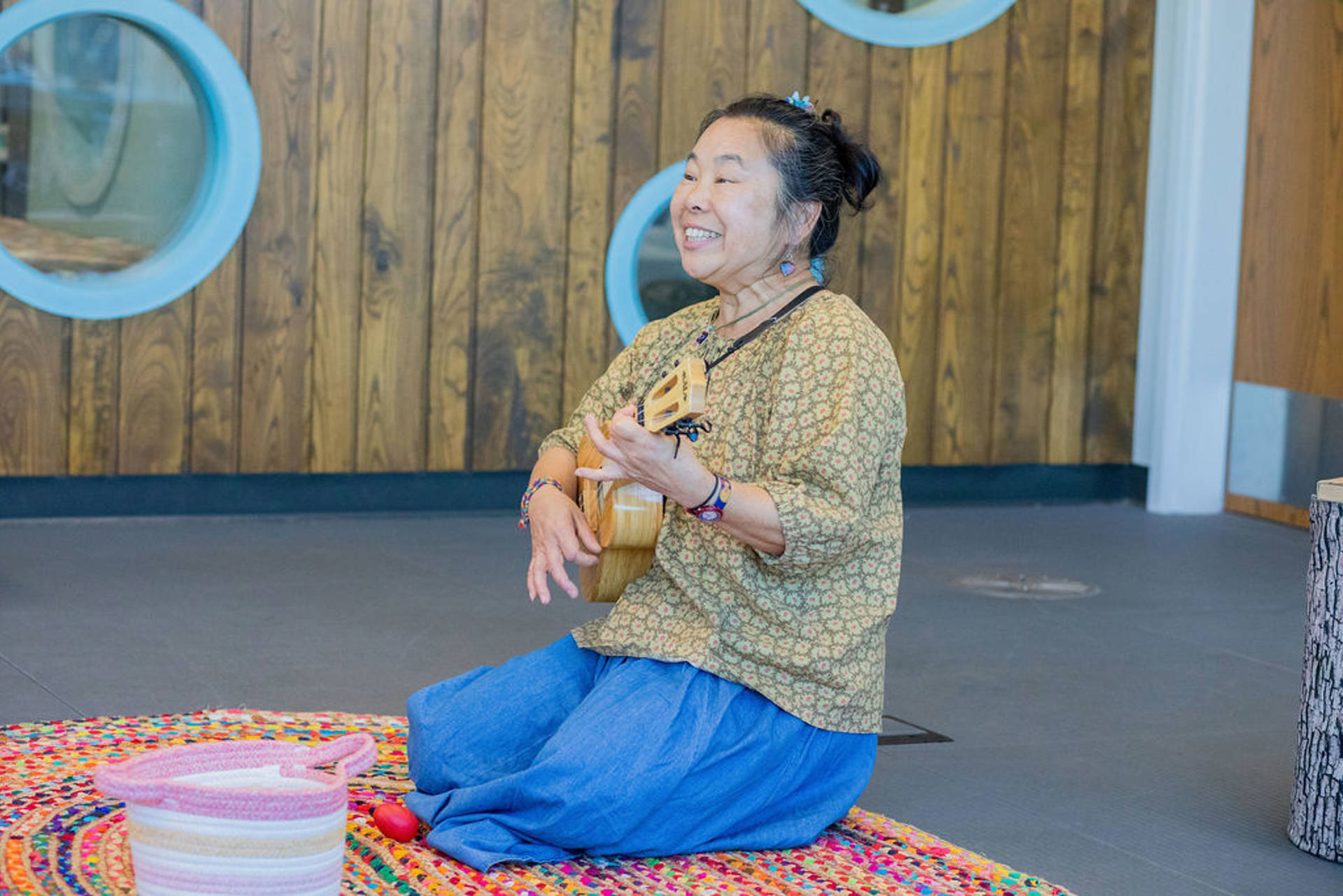A woman with medium complexion and dark hair sitting on the ground playing a ukelele. She is smiling.