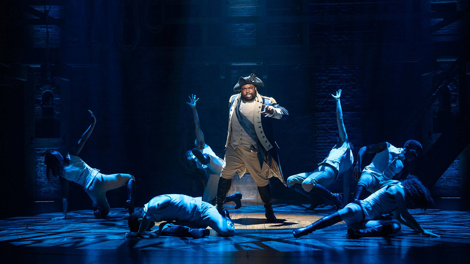 A man with a dark complexion wearing a triangular hat and blue coat sings beneath a spotlight while surrounded by dancers.