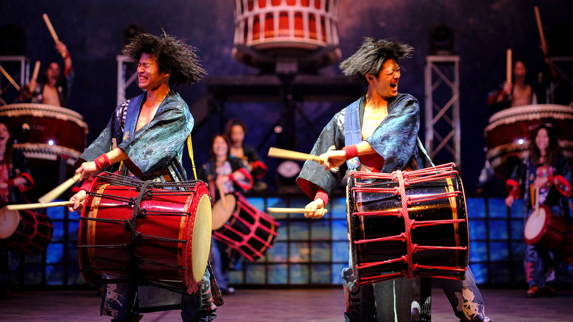 Two Japanese men with cropped black hair intensely playing drums strapped across their chests in unison. They are both wearing blue kimono robes.