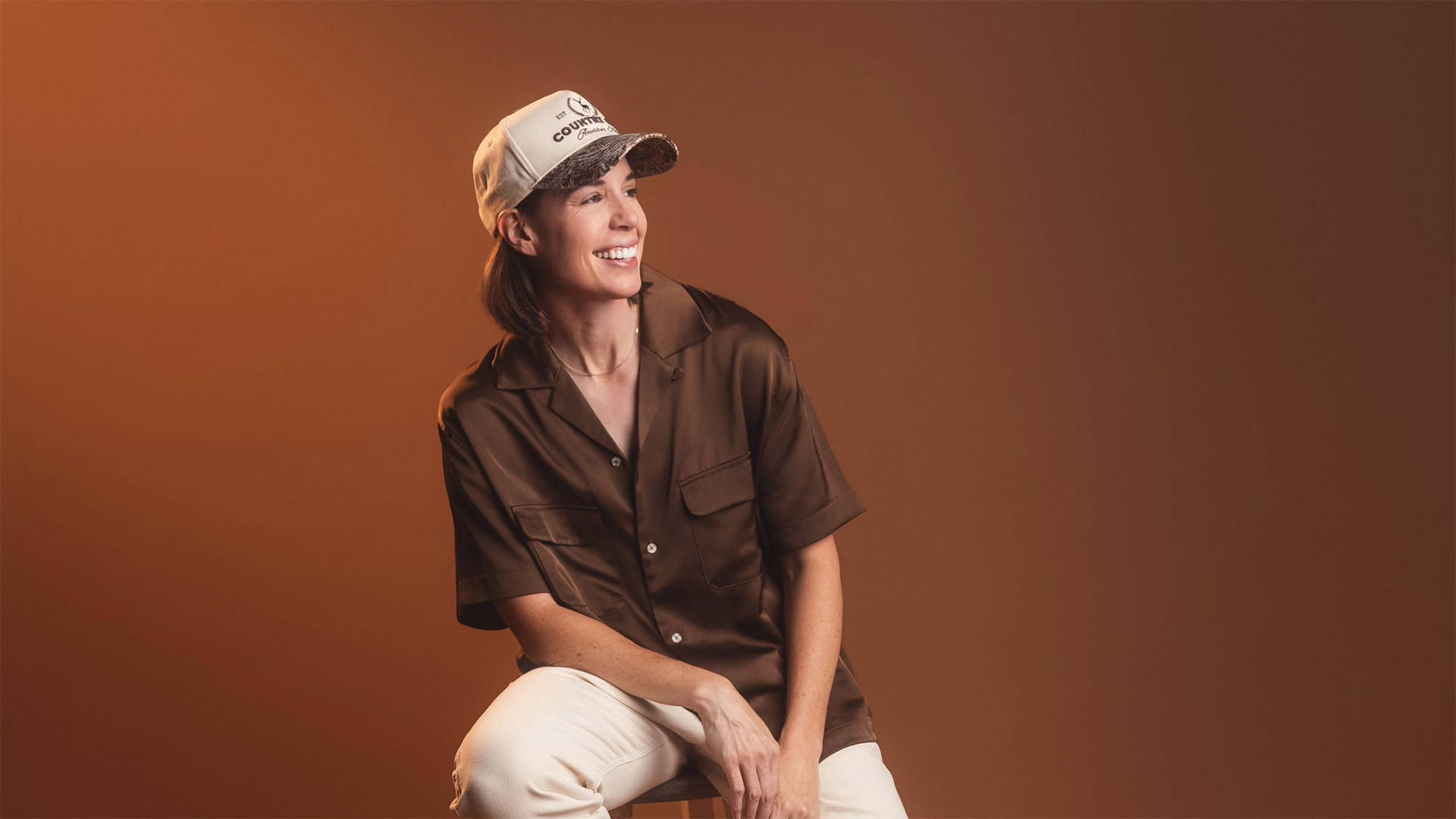 Danae Hays, a woman with light complexion, medium length brown hair, a trucker cap and a baggy brown button up shirt, sitting on a stool and smiling.
