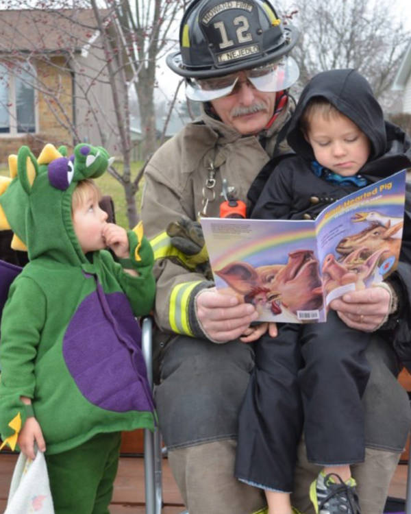 A firefighter with a moustache reading a book to two small children in cute halloween costumes.