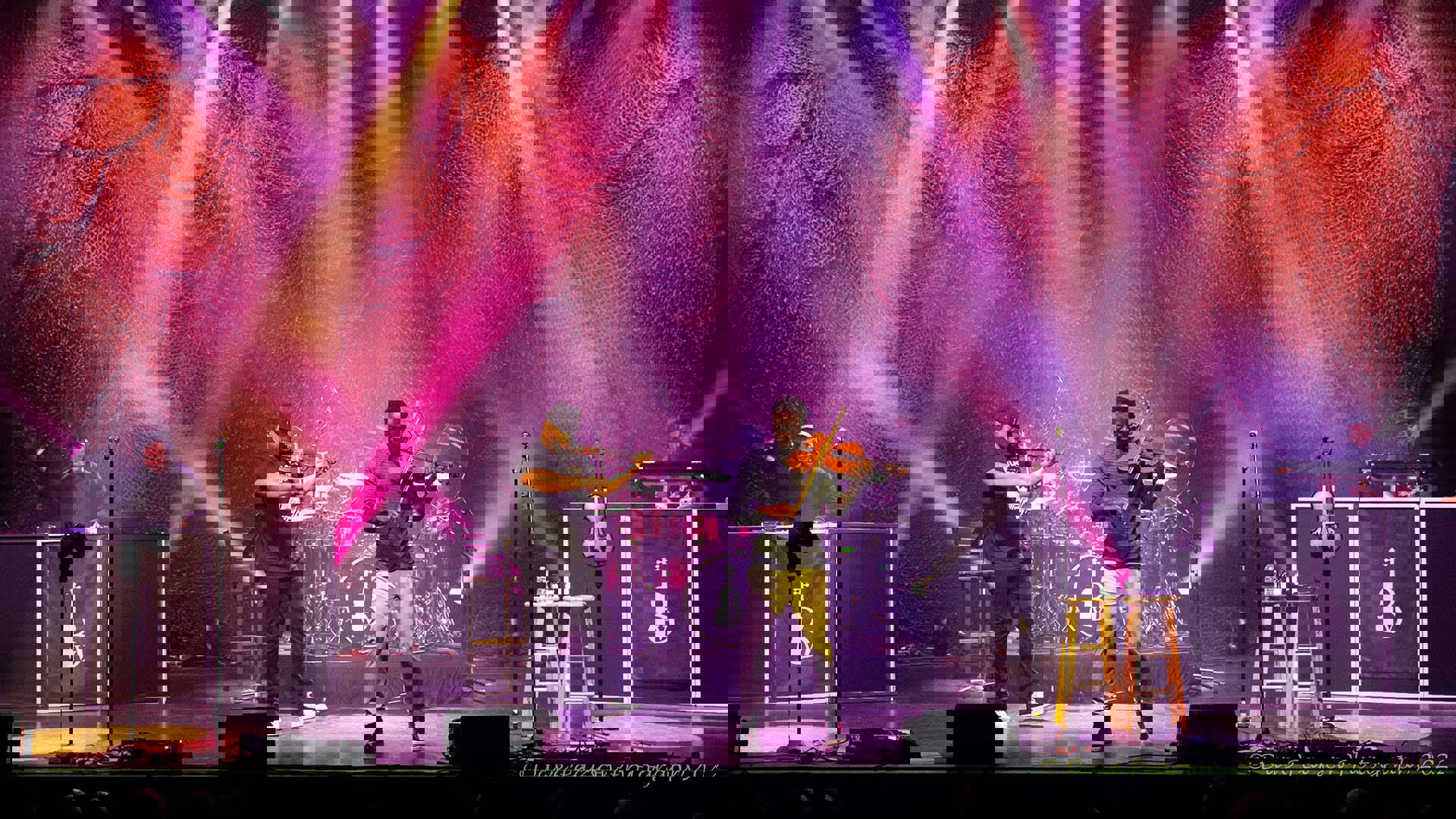 Black Violin, two black men playing violin and viola in casual clothes, performing on stage with pink and orange stage lights behind them.