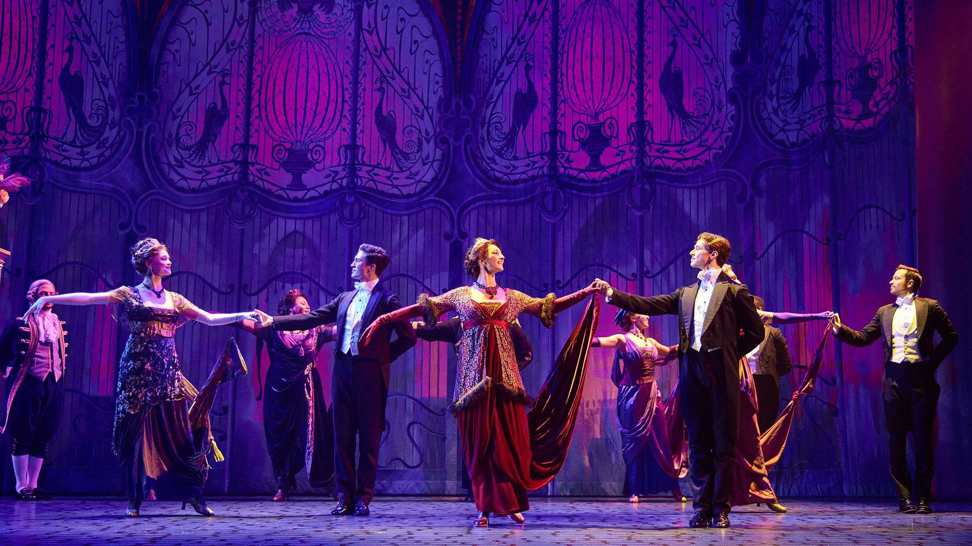 A group of couple dance in from of a pink and purple background. They are dresses in fancy party dresses and black and white tuxedos.
