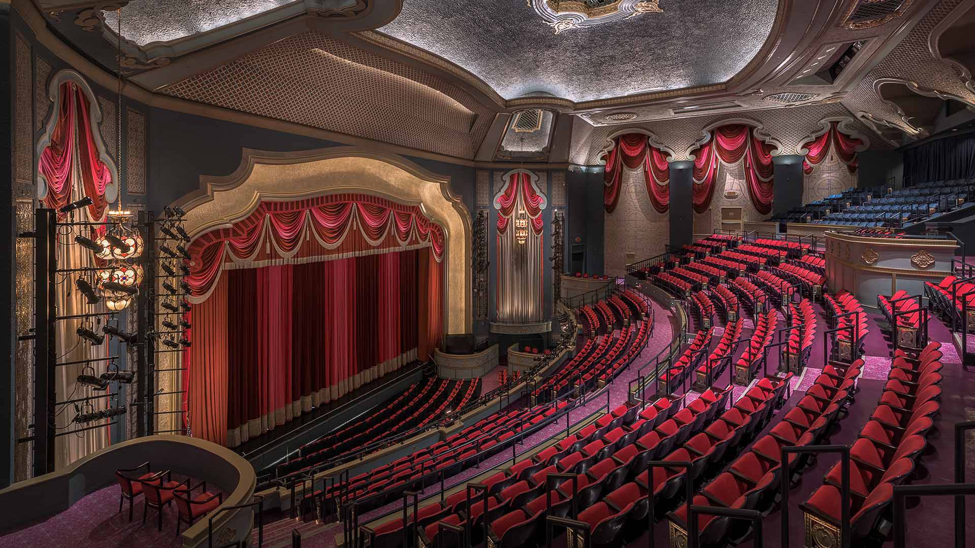 A view from the balcony of Capitol Theater looking down to a stage with a red curtain. The empty chairs are also red.