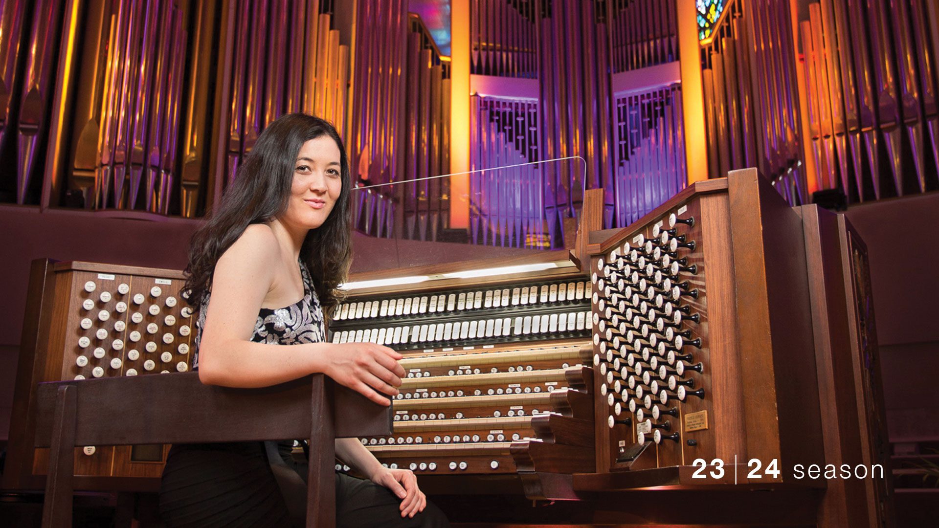 A young woman with light complexion and long dark hair is sitting at an organ console. She is turned around and looking back at the viewer.