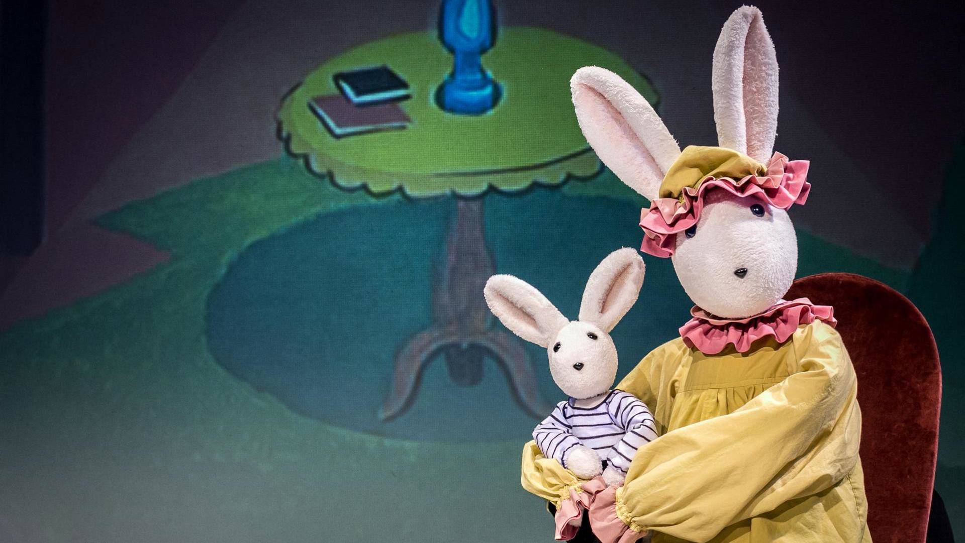 A large bunny puppet in a yellow dress holding a smaller white bunny puppet wearing a white & blue striped shirt. They are sitting on a red velvet chair.