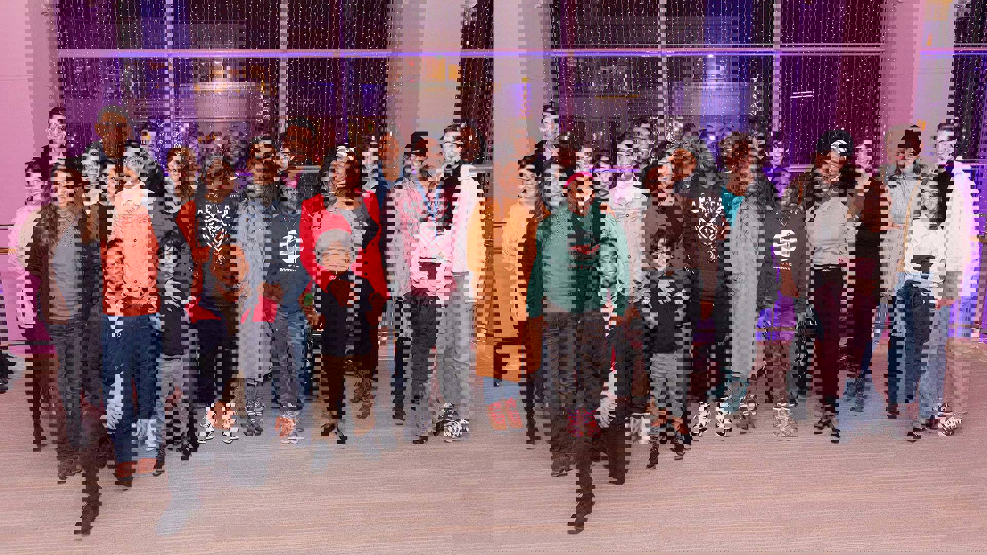 A group of smiling adults and children posing together in front of large windows and white columns with thin white vertical lights