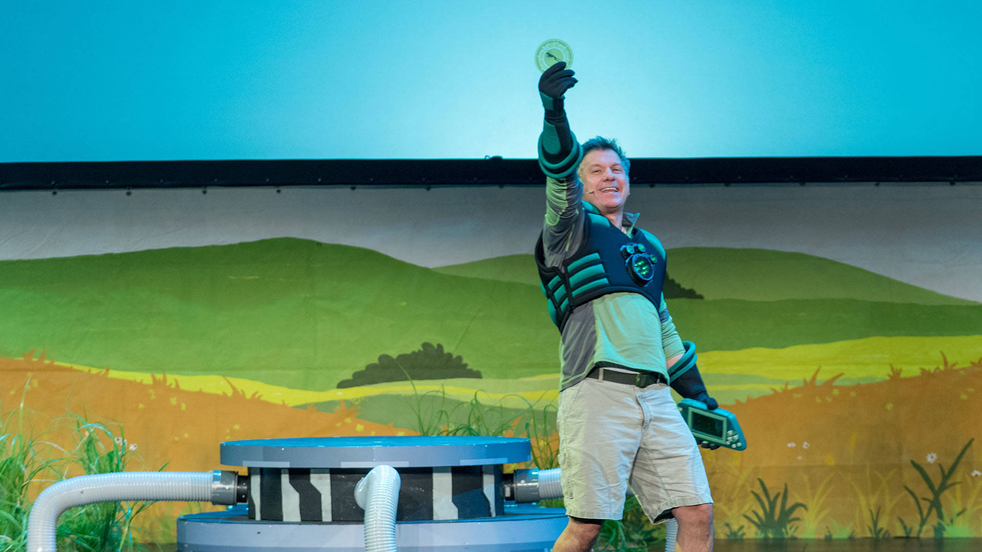 Chris Kratt, a man with light complexion wearing shorts and superhero vest/gloves, holds up a green disc during a LIVE 2.0 performance.