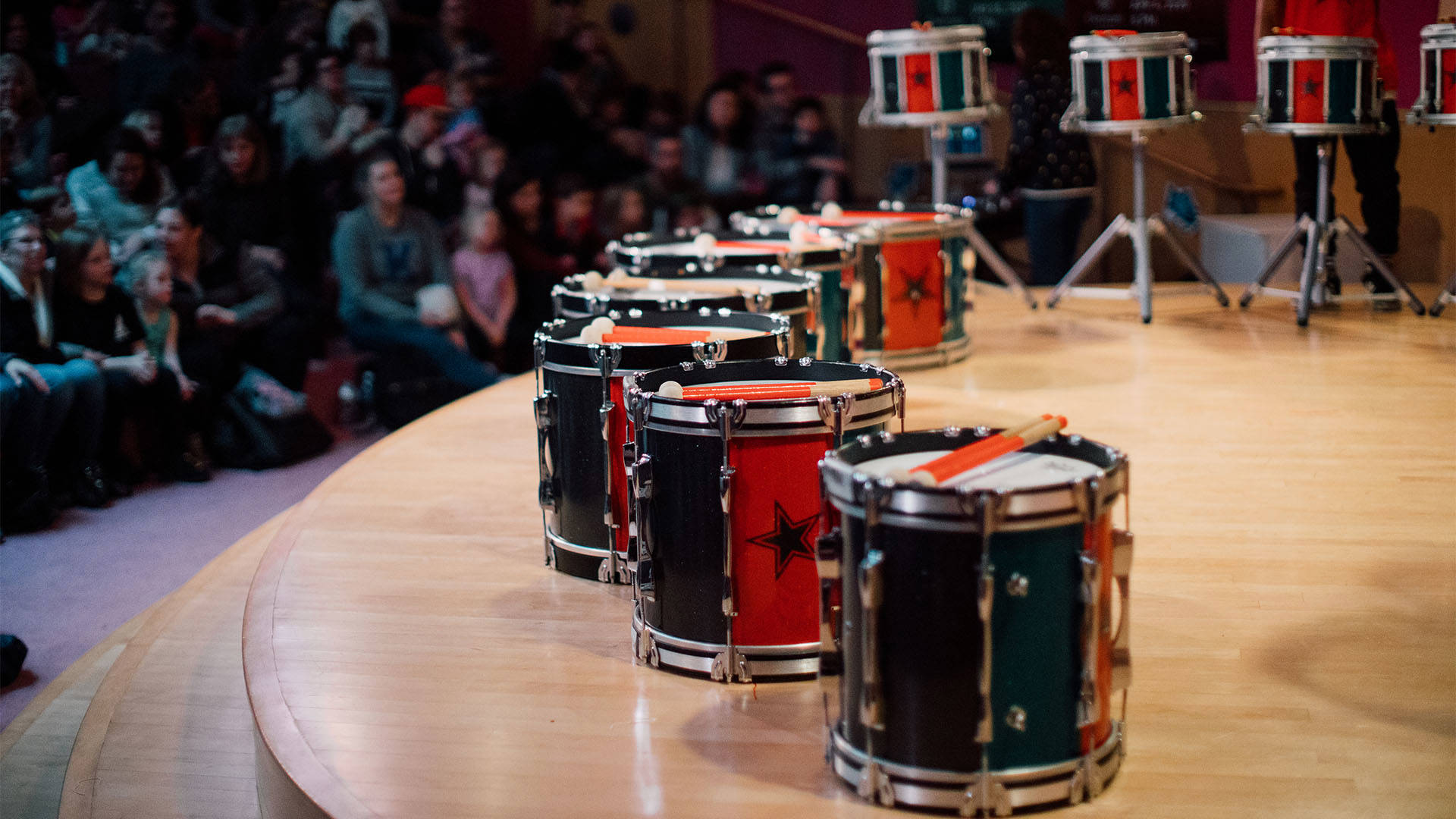A line of black and red marching bass drums on a wooden stage. Marching snare drums are on stands in the background