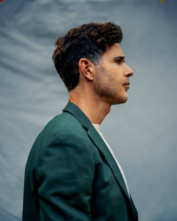Profile view of a young man with light complexion, short dark hair, and a teal suit with a blue photography background behind him.