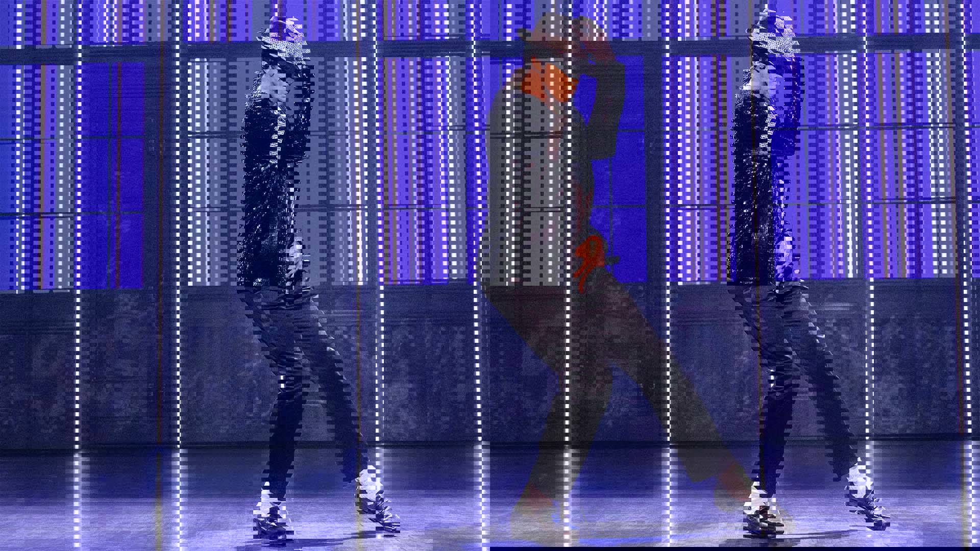An actor playing Michael Jackson dancing on stage. He has medium complexion, dark hair, a white shirt, dark sparkling jacket, and dark pants. The background is purple.