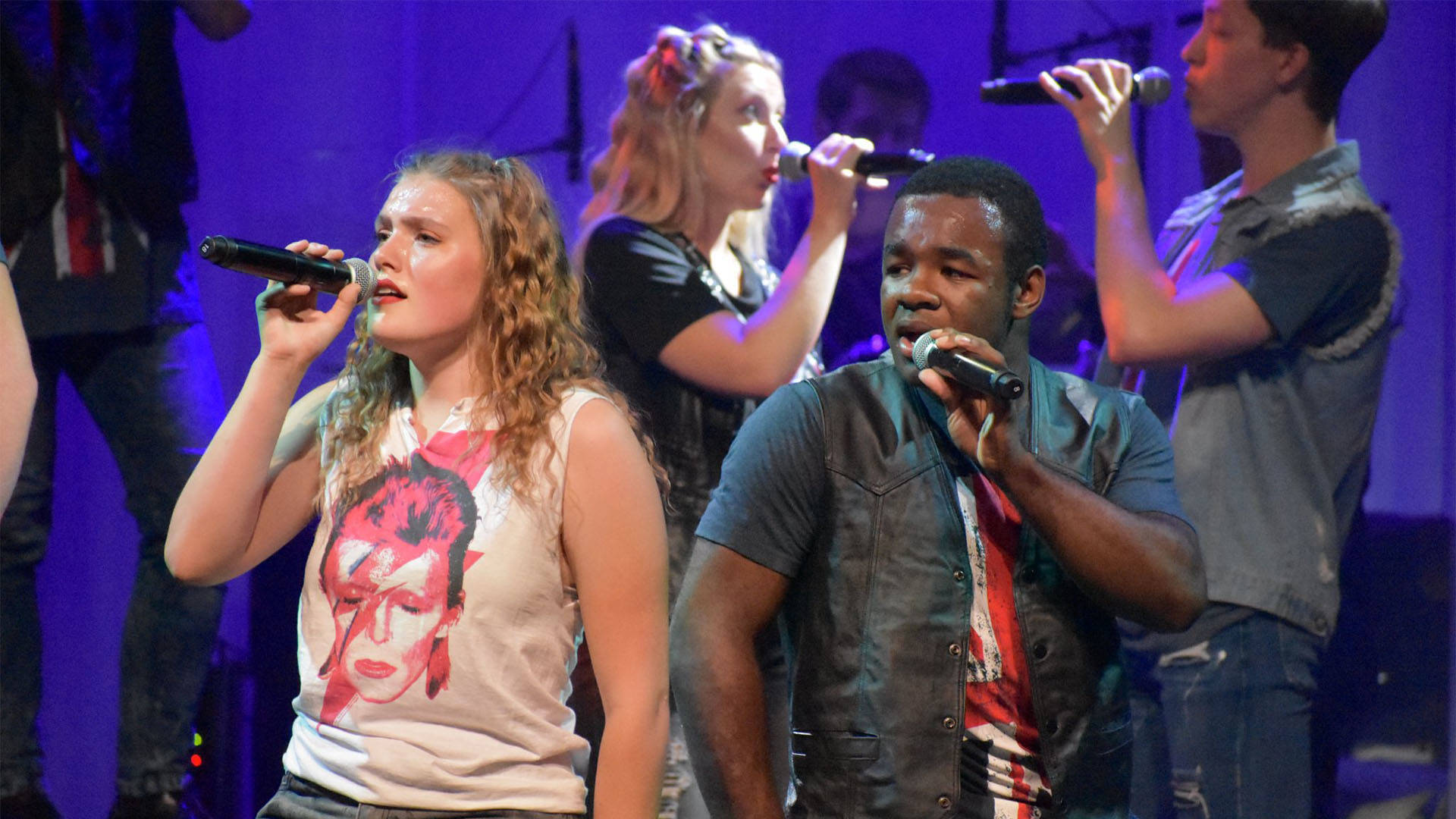 Four high school aged kids singing into microphones against a purple background.