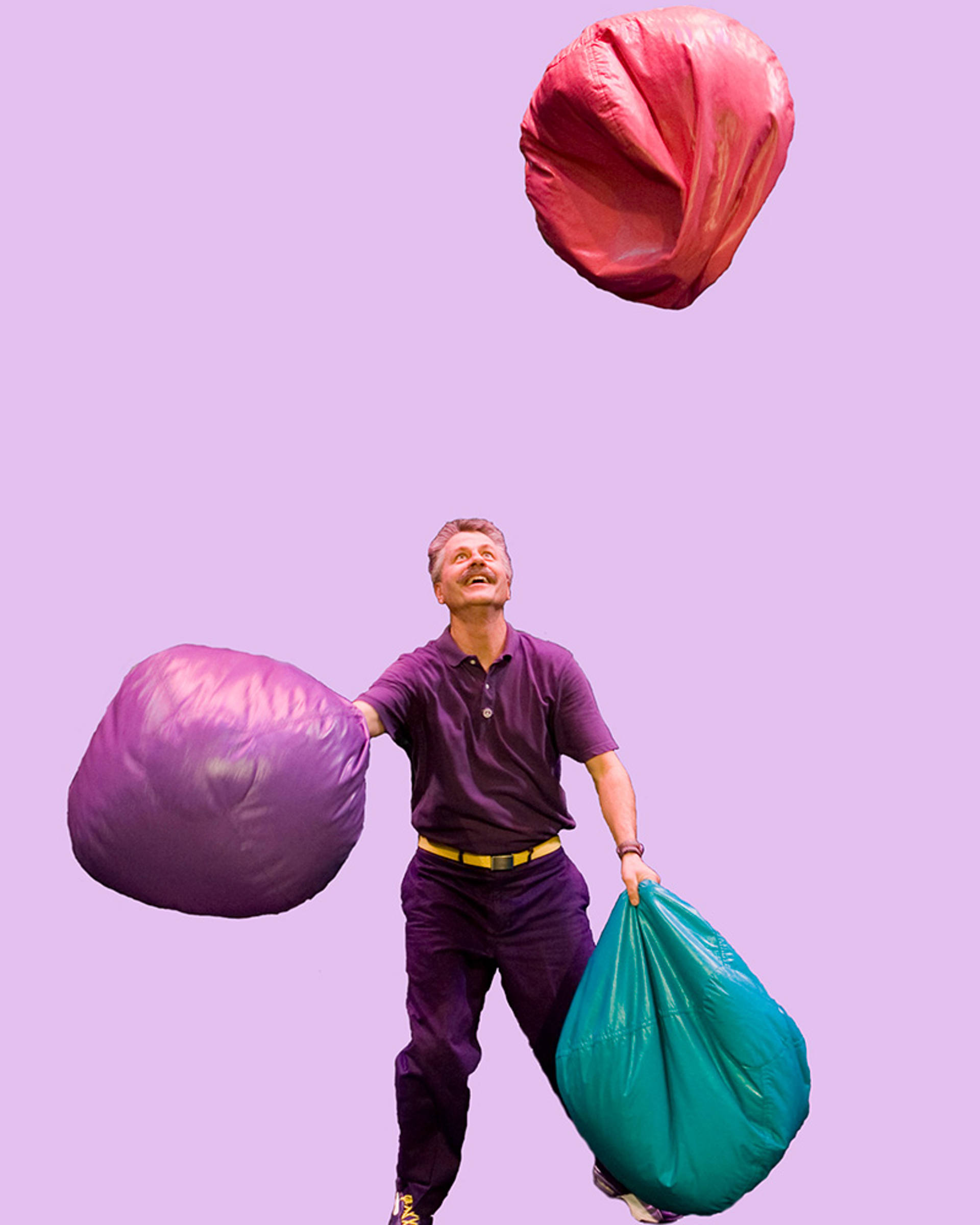 A man dressed in all purple with short grey hair and a moustache throughing colorful bean bags into the air.
