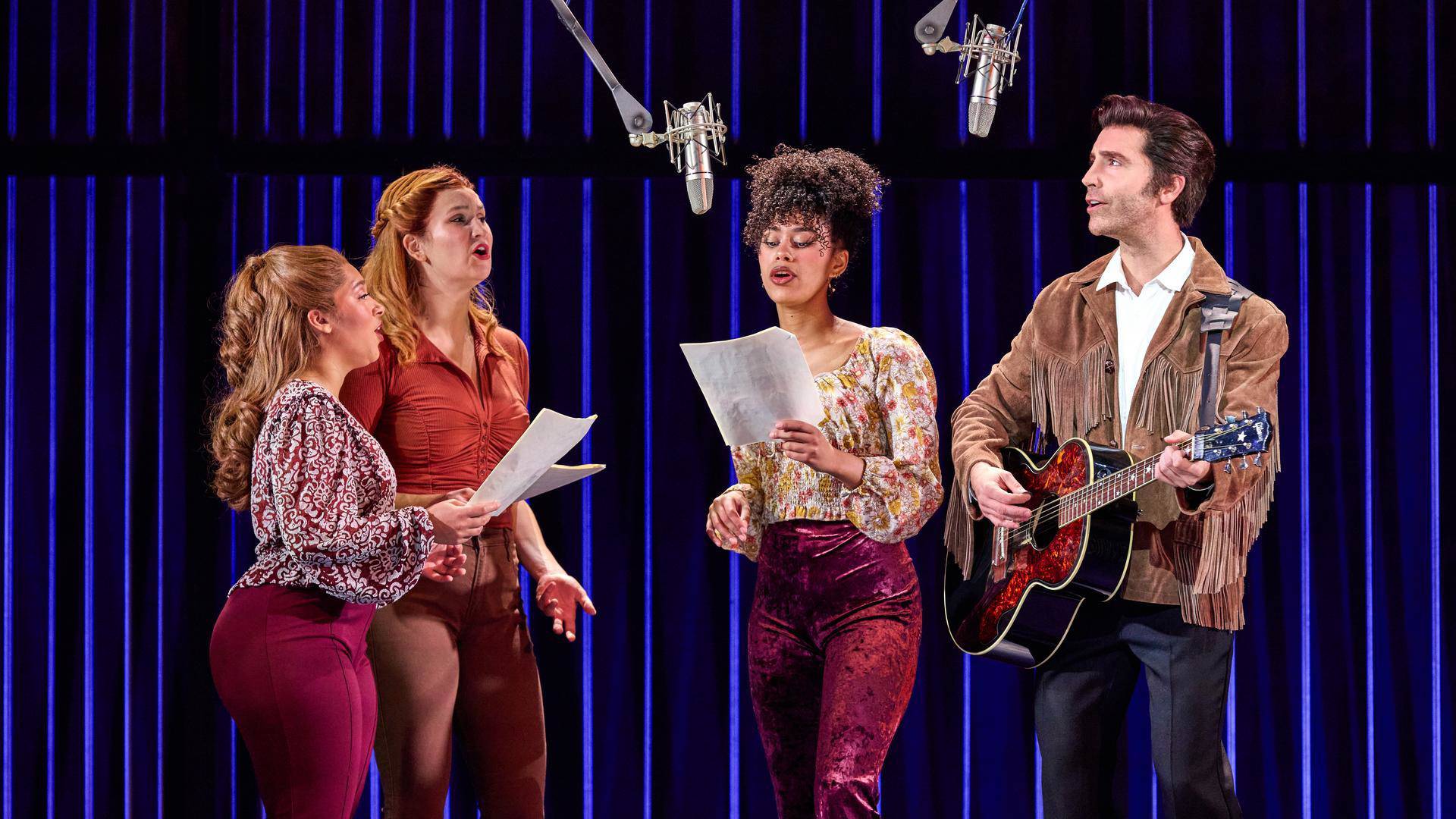 Four peopl huddling around suspended microphones and holding lyric sheets. On the right a man holdings an acoustic guitar.