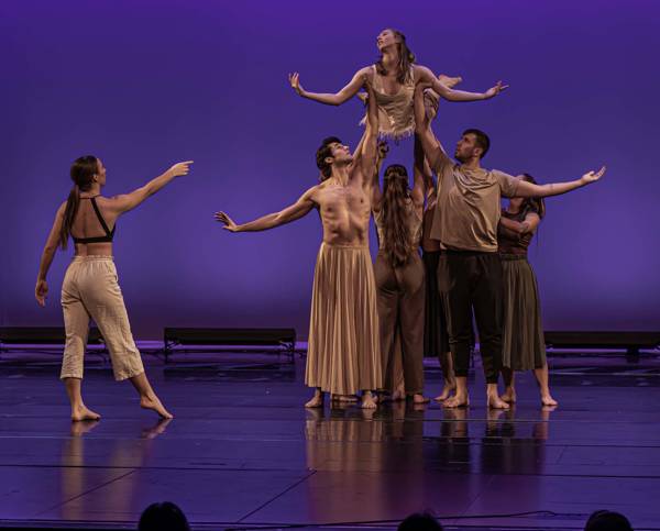 Dancers on a stage with a purple backdrop. They are wearing various tan outfits. Five dancers are holding one dancer above their heads while one dancer stands to the left and points at them.