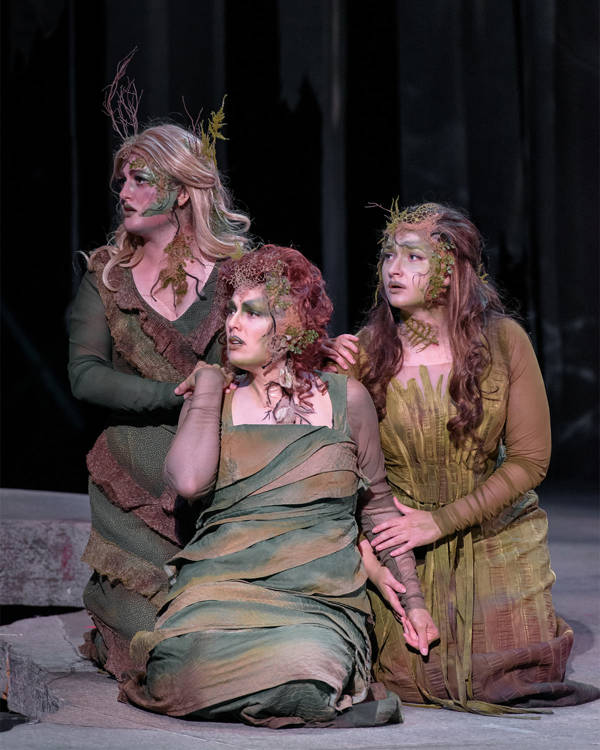 Three caucasian women with concerned looks on their faces kneeling together. They are each wearing earthy green costumes and facepaint.