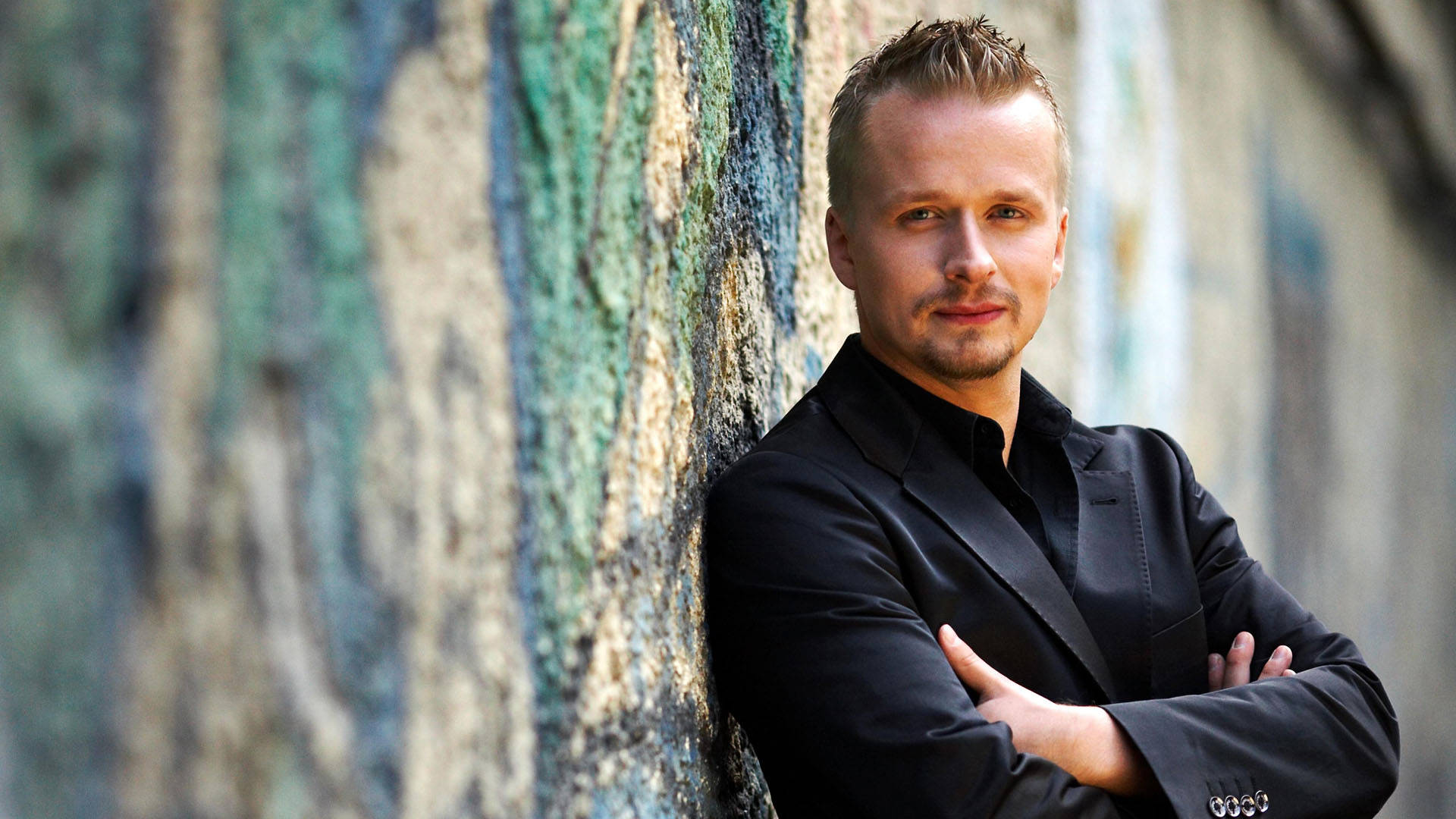 A white man with short blond hair and a goatee standing against a cement wall painted white, blue and teal. He is wearing a black suit and shirt with his arms crossed, looking at the camera.