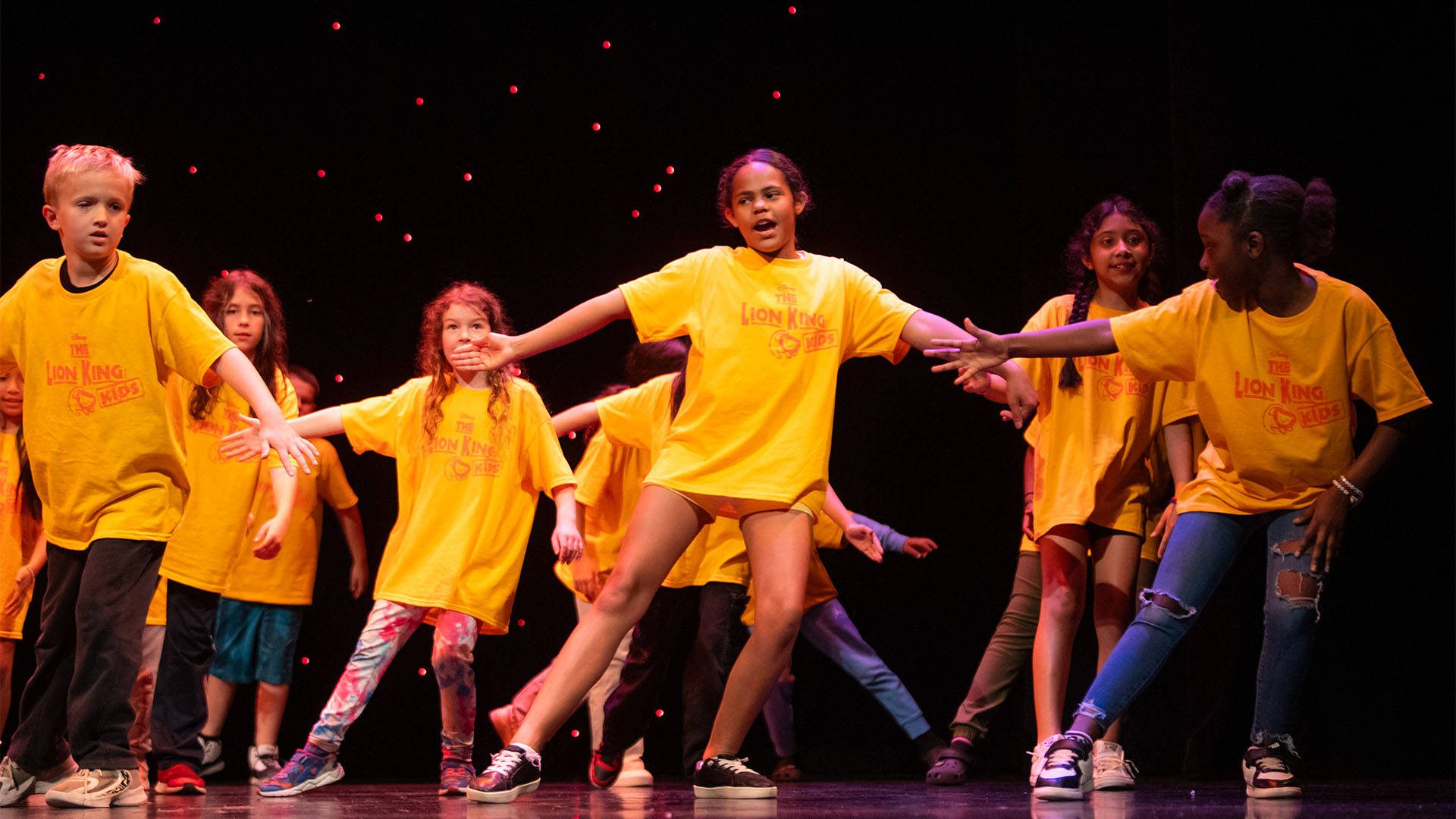 A diverse group of young students singing and dancing on stage in yellow "The Lion King" t-shirts.