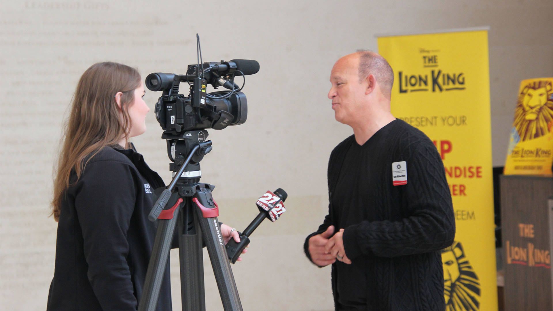 A woman with brown hair next to a news camera filming an interview with a man in black with short hair.