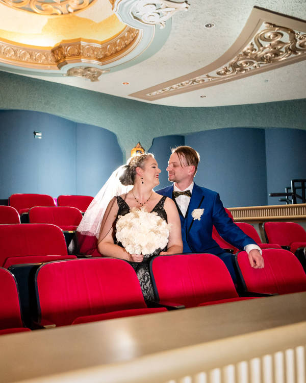 A man in a blue suit and a woman in a black dress sitting in red Capitol Theater seats and looking at eachother.