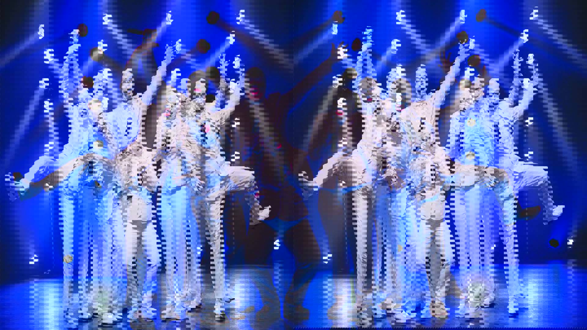 Eight performers strike zany poses against blue lights. They all have full white suits on and full white facepaint and baldcaps.