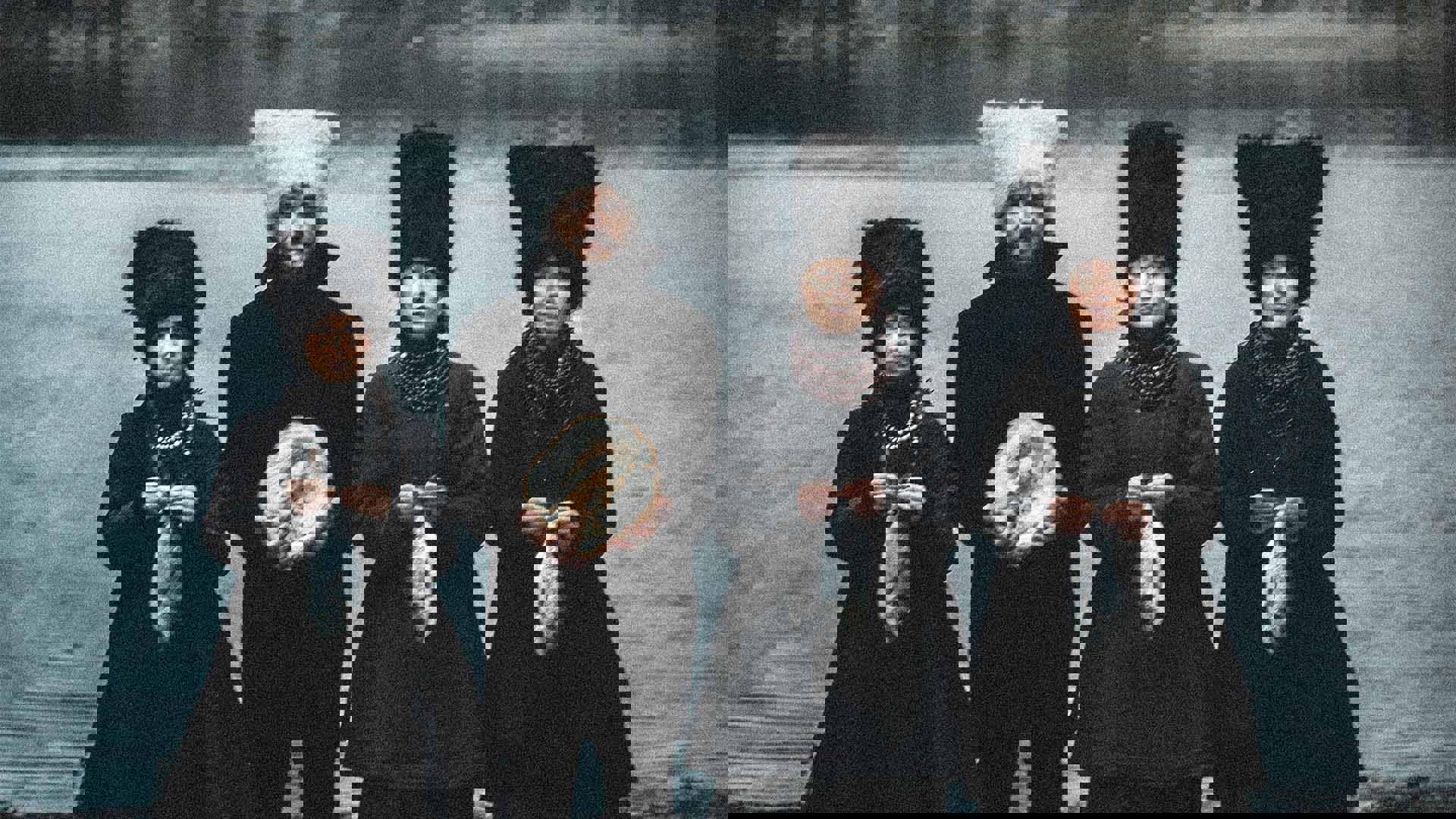 Four people with light complexion stand holding fish. They are wearing ornate traditional black Ukrainian outfits. Three have large plush black headwear on and the one man has a white hat on 