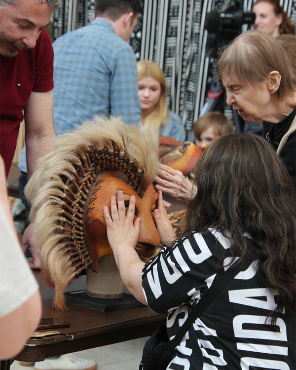A wooden lion mask on a table. People are standing around it and touching it.