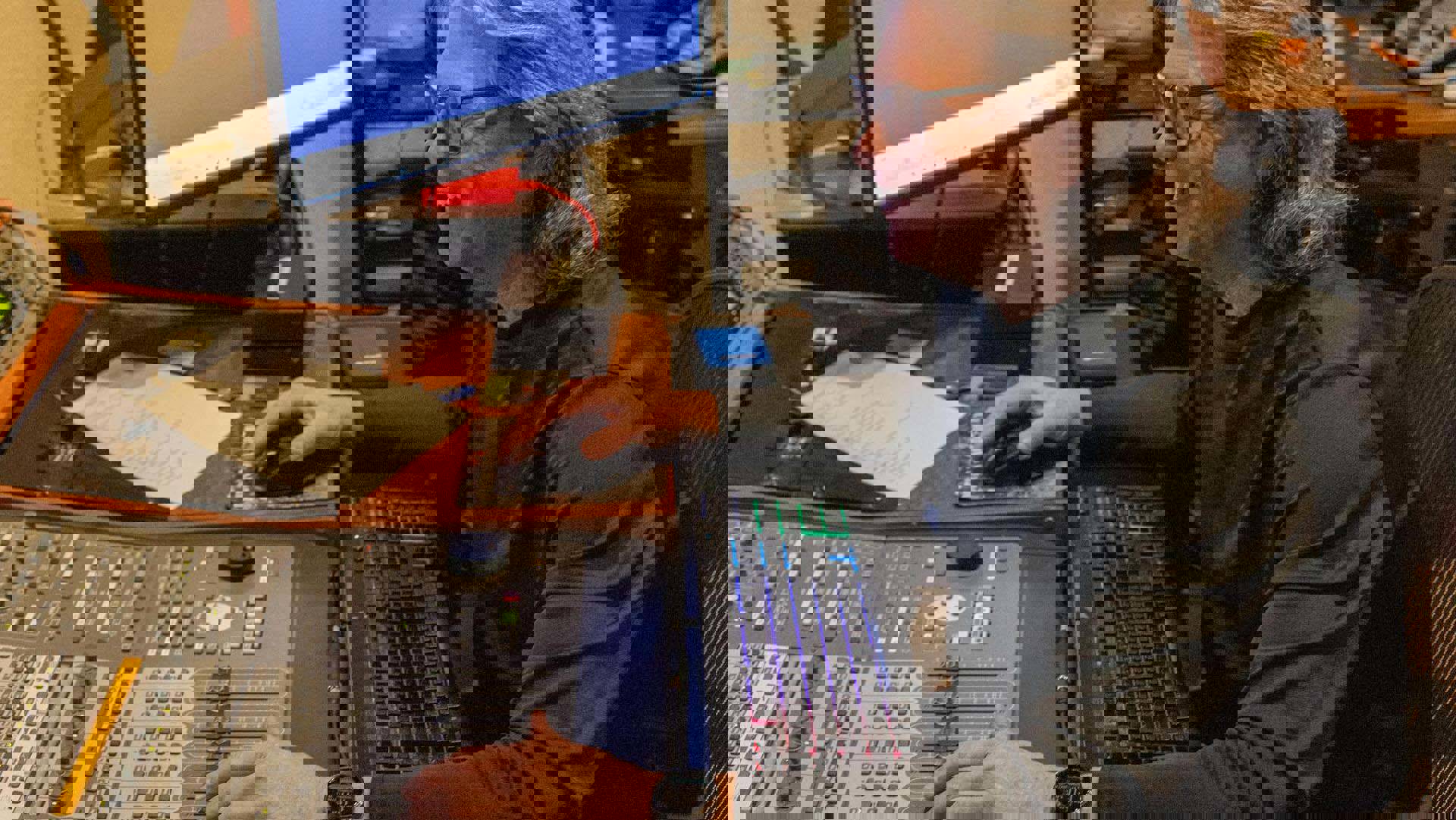 A man with light complexion and long grey hair with glasses in a black shirt sitting at a recording studio control console.