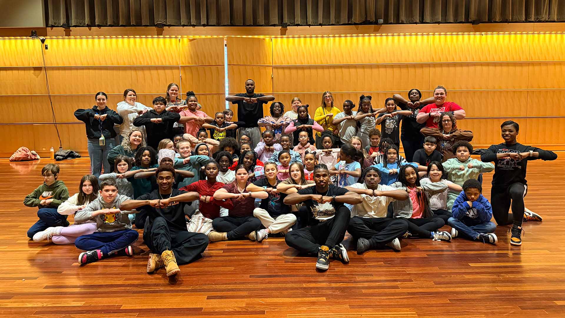 A group of elementary schoolers hold their fists together with their elbows up while posing for a picture on a wood floor.