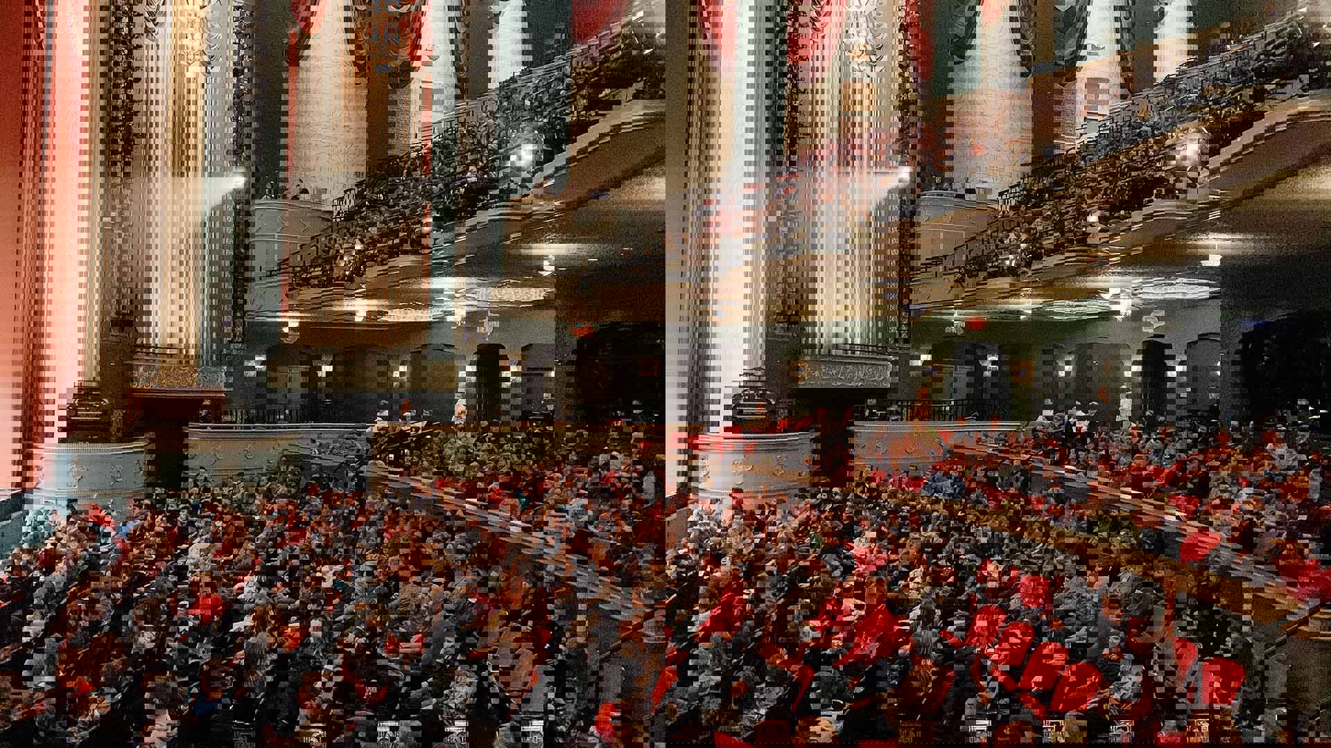An audience sitting in Capitol Theater before a show. The theater has red seats, white and green walls, and red curtains.