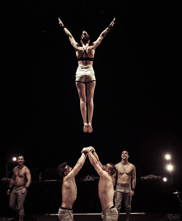 A woman in the middle of a jump. Her back is facing the camera. Two shirtless men stand below ready to catch her.