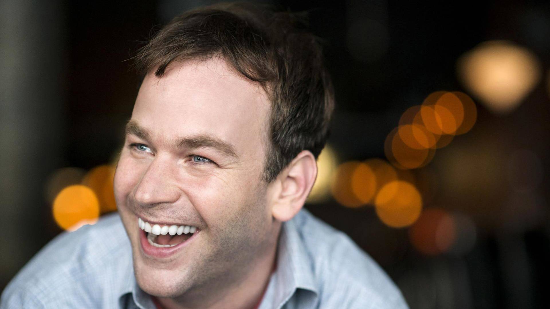 Headshot of Mike Birbiglia laughing . He is a caucasian man with short brown hair and blue eyes. He is wearing a light blue collared shirt.