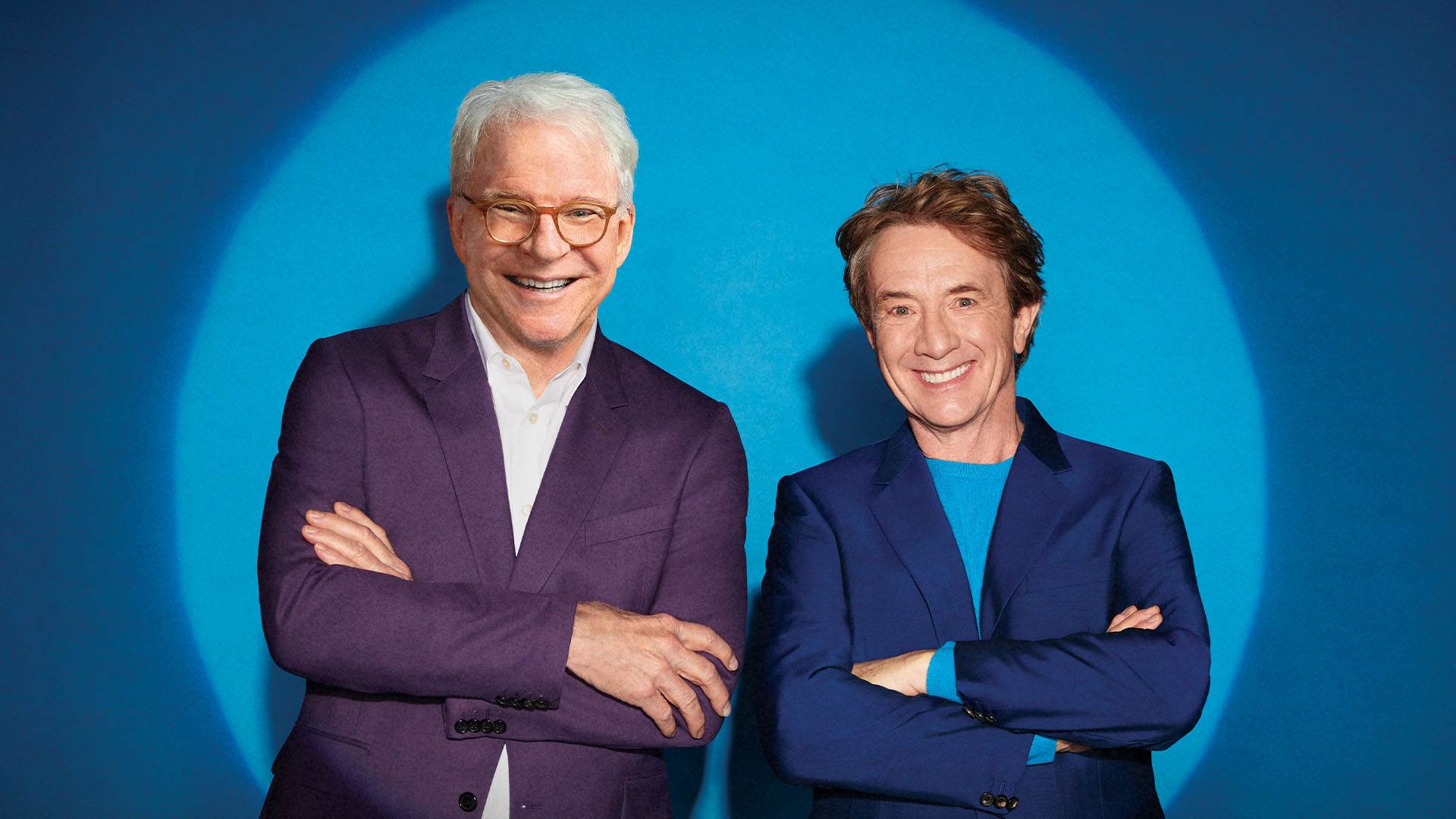 Steve Martin & Martin Short stand together with their arms crossed against a blue wall illuminated by a spot light. They are both caucasian men. Steve Martin, on the left, has white hair, glasses and a plum colored jacket. Martin Short, on the right, has reddish brown hair and a blue jacket.