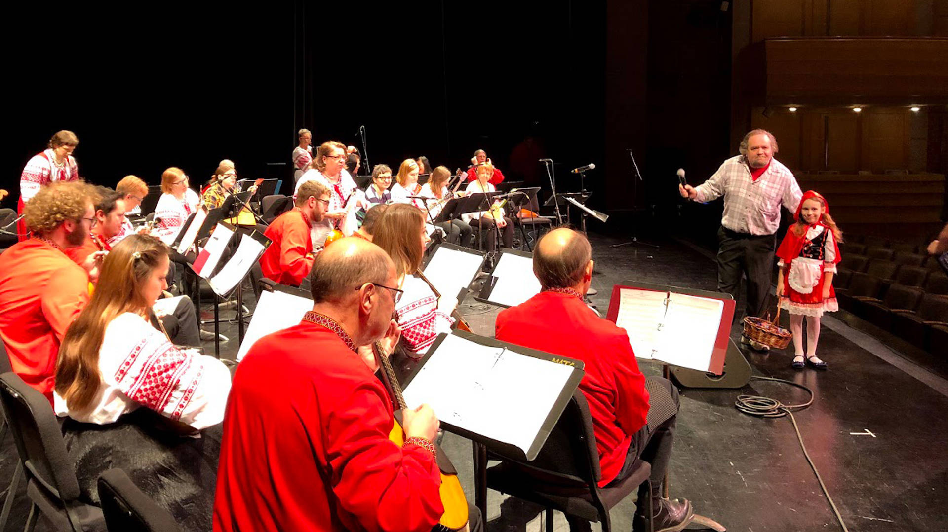 A russian folk orchestra rehearsing on a dark stage. The members are wearing red and white outfits. on the right a conductor and a young girl are looking towards the camera.
