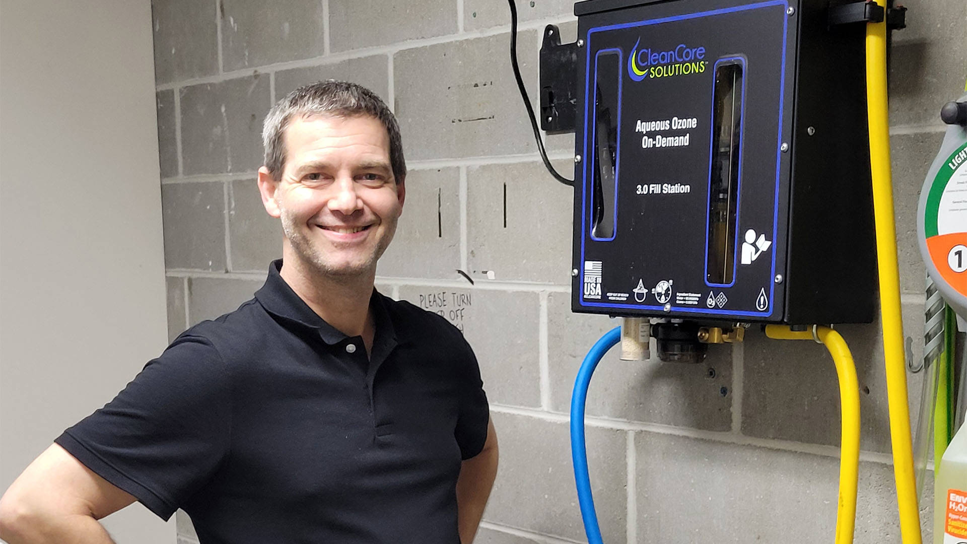 A middle aged caucasian man with short hair and a dark polo standing next to fill station equipment hanging on a cement wall