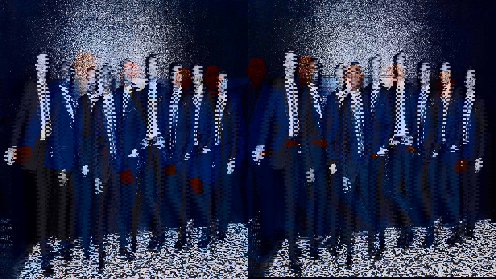 A group of smiling men in blue suits standing against a black wall.