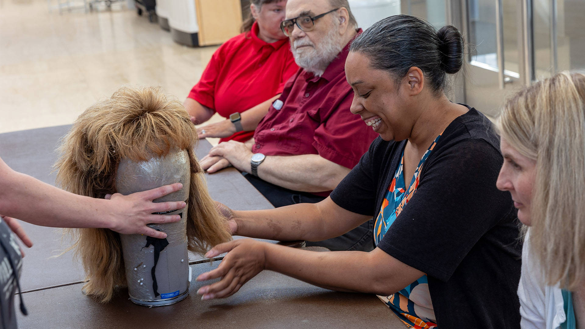 A black woman with black and grey hair in a bun is smiling and lightly touching a Tina Turner 80s mullet inspired blonde wig at a table with others.