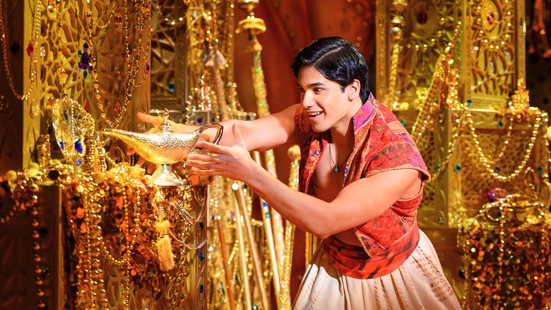 A man with short, dark hair standing in a room filled with gold. He is holding a gold lamp and rubbing it. He is wearing an orange vest.
