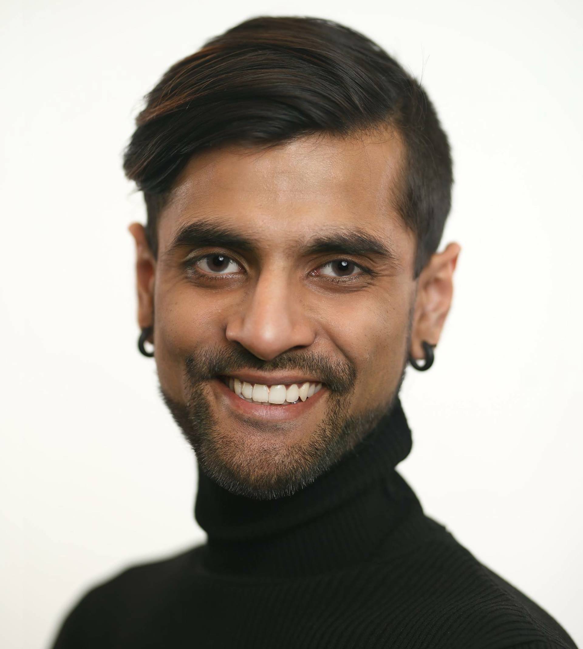 A young man with medium skin tone, dark hair, a beard, and earings in a dark turtleneck top.