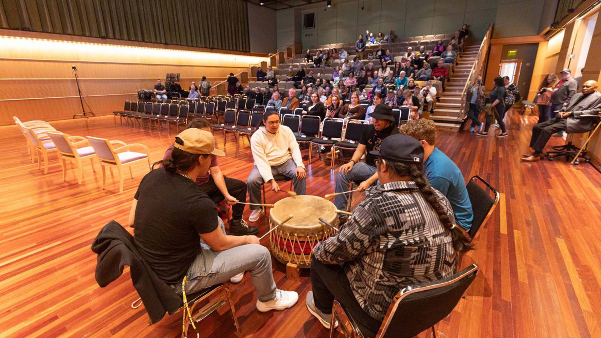 Six people with medium and light complexion sitting in a circle of chairs surrounding a large animal-hide drum. Each person is holding a long mallet and hitting the drum in synchrony. They are in Promenade Hall at Overture Center with an audience behind them.
