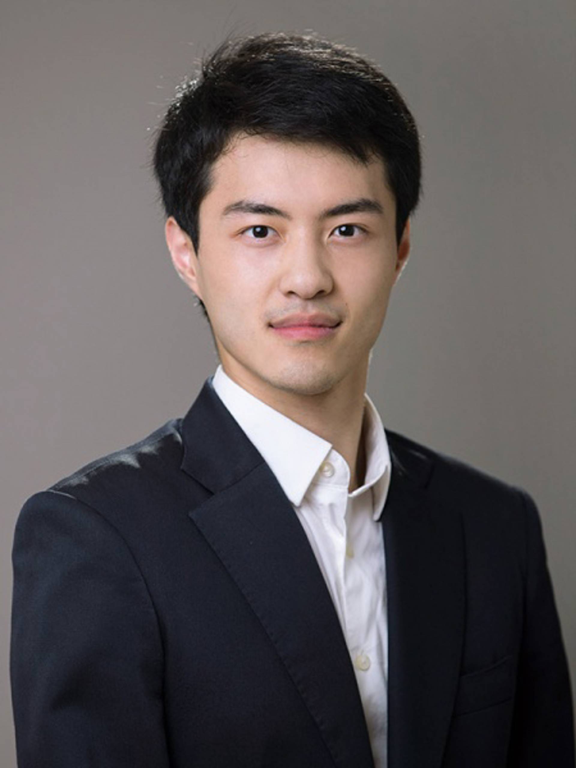 Headshot of a young man with medium complexion, short black hair, a white collared shirt and a dark jacket against a grey background