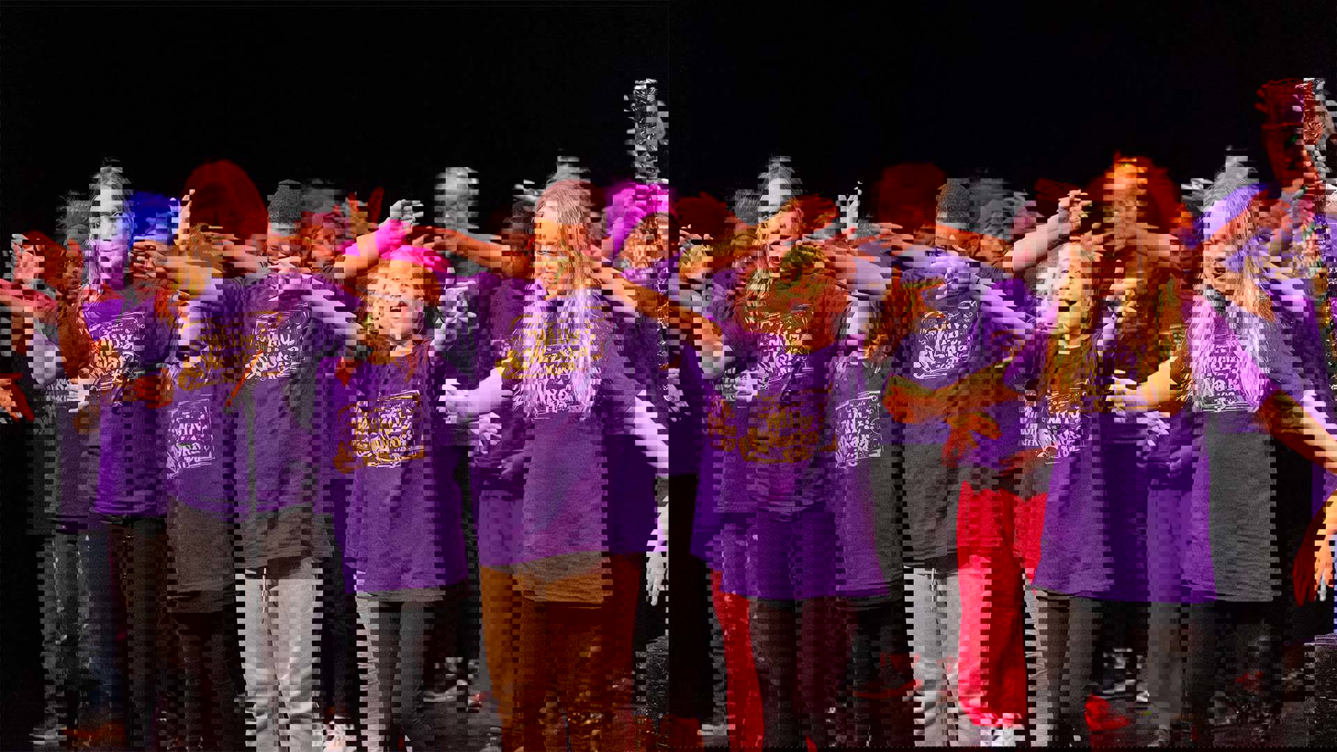 Young students in purple shirts and colorful headwear performing on a dark stage. 