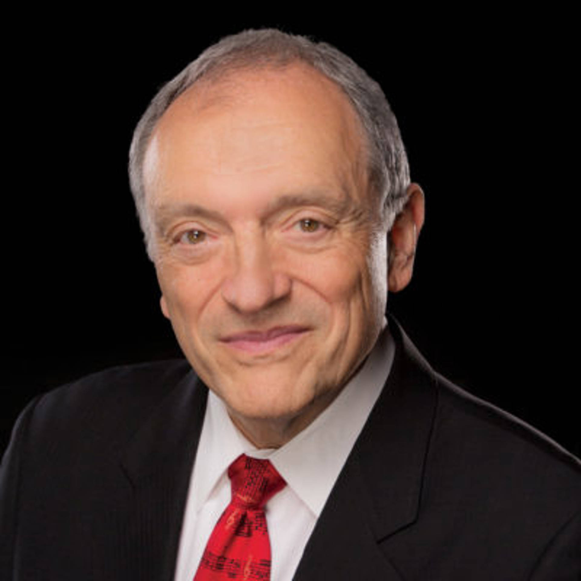 Headshot of an older man with light complexion, short grey receding hair and brown eyes. he is wearing a dark suit with a red tie.