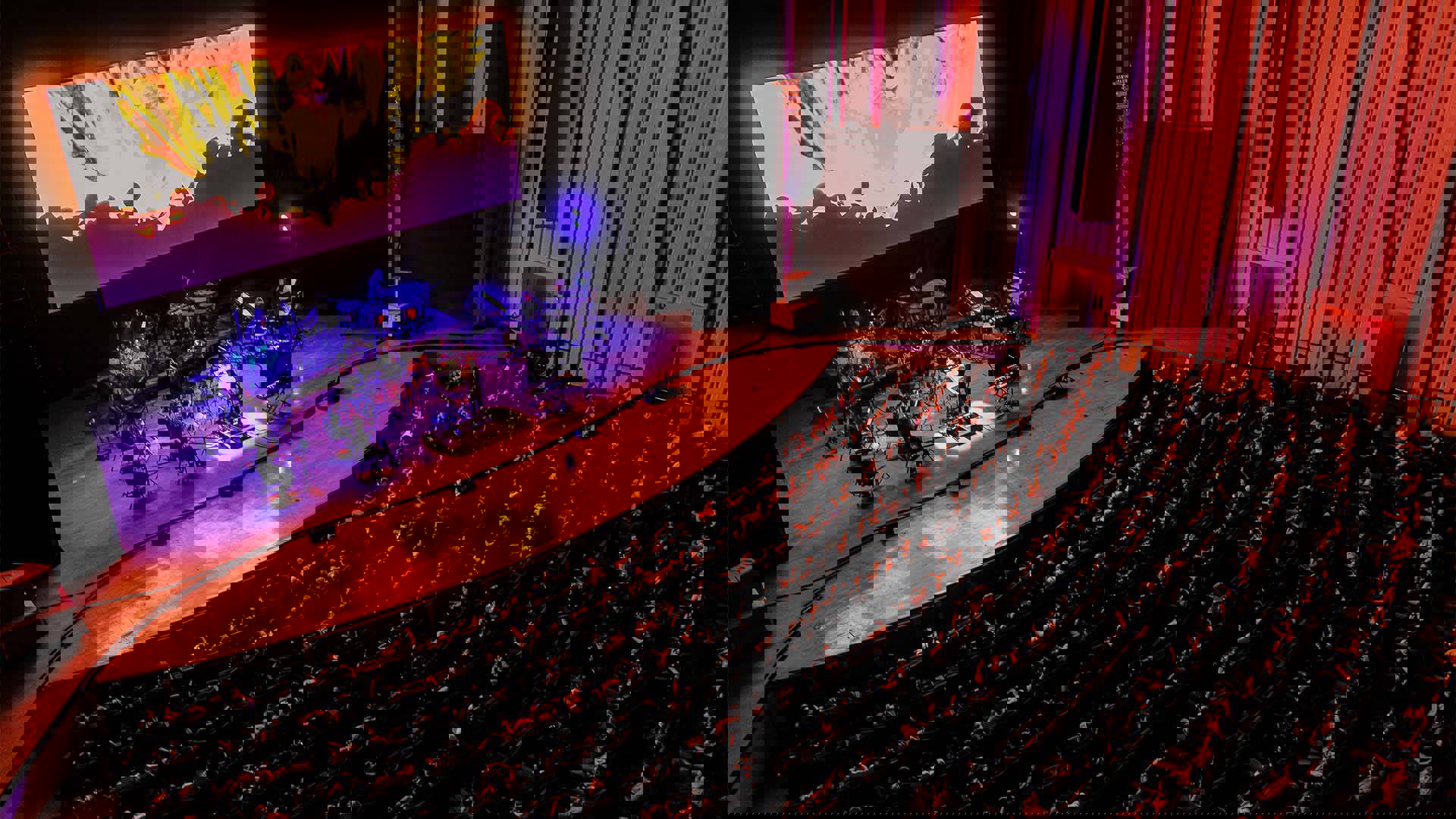 A full house audience watching an orchestra performing on stage under a large screen with a scene from Spiderman into the Spider-verse.