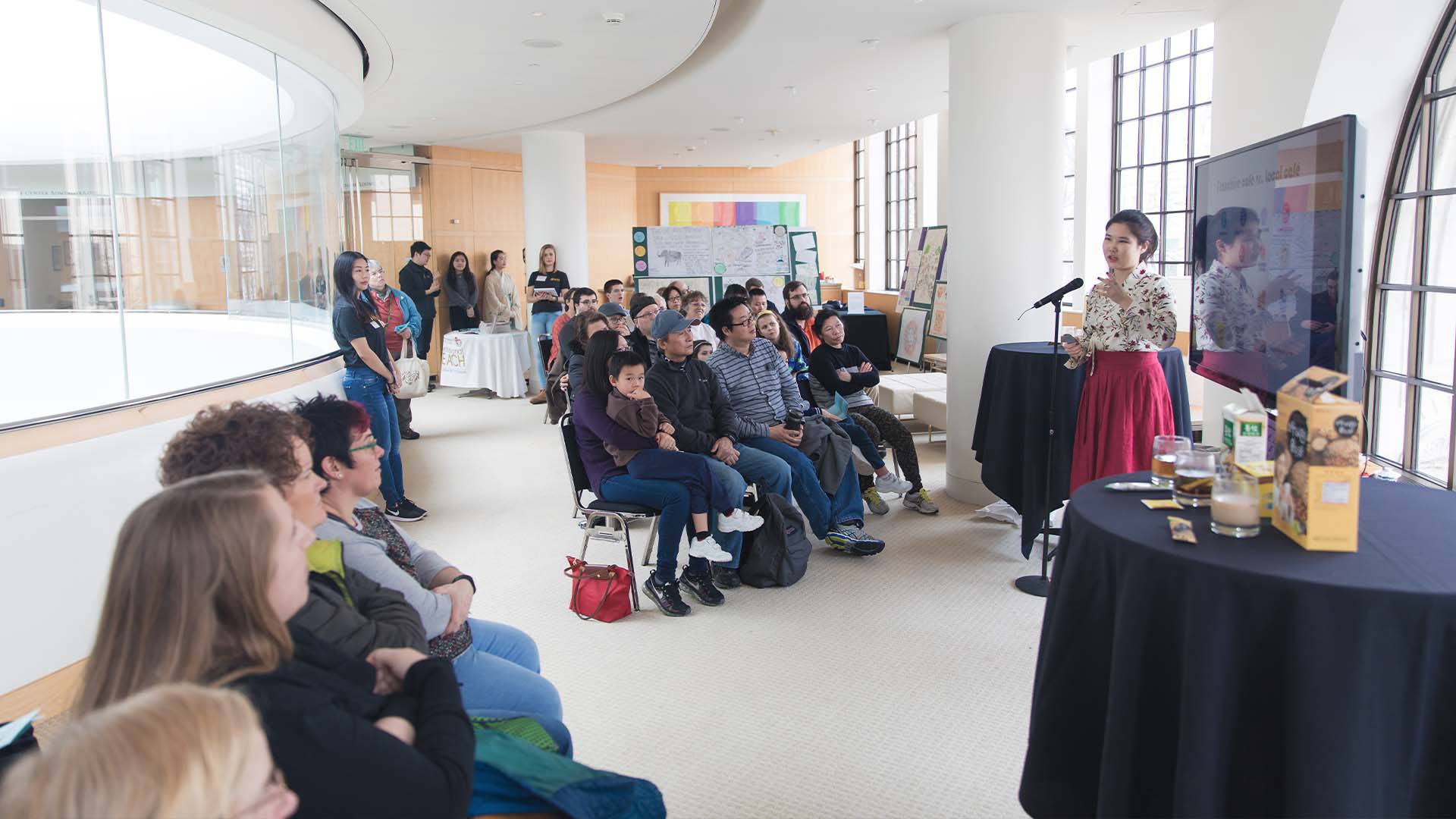 A group of people watch a presentation. The room is long and curved.