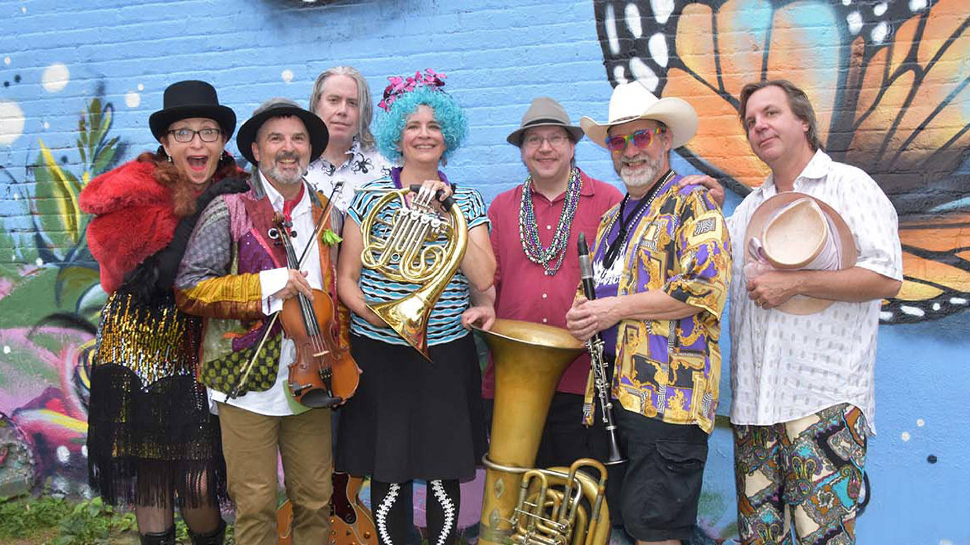 Seven Yid Vicious band members in bright and colorful clothing stand together and smile. They are holding instruments including violin, french horn, tuba and clarinet.