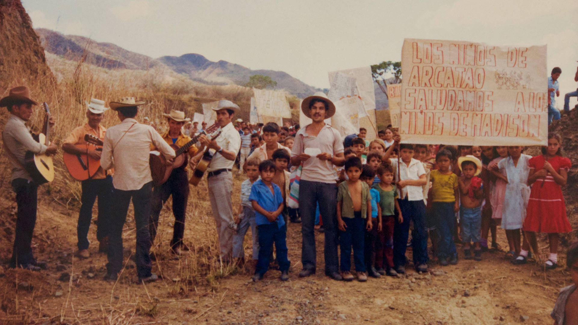 Photo of a group of people standing outside posing for a photo with signs in spanish. The most prominent reads" Los ninos de Arcatao saludamos a los ninos de Madison"  On the left five men hold guitars and are playing together.