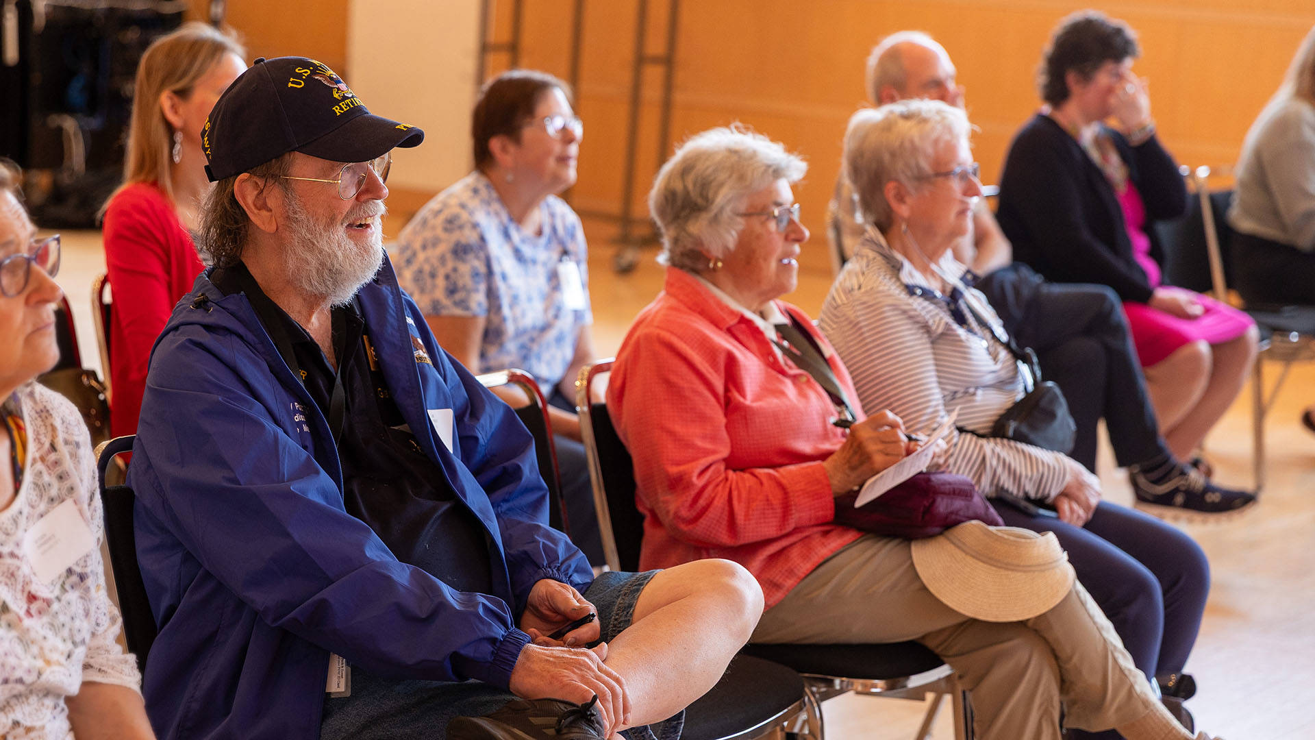 Two rows of elderly adults in casual clothing sitting watching a performance in a bright lit room.