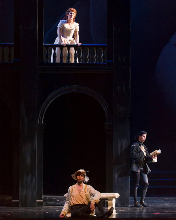 Performers in the opera "Don Giovanni" performing on a stage. A woman in a white dress is on a balcony above a seated man in peasant's clothing and a man in fin dark clothing.