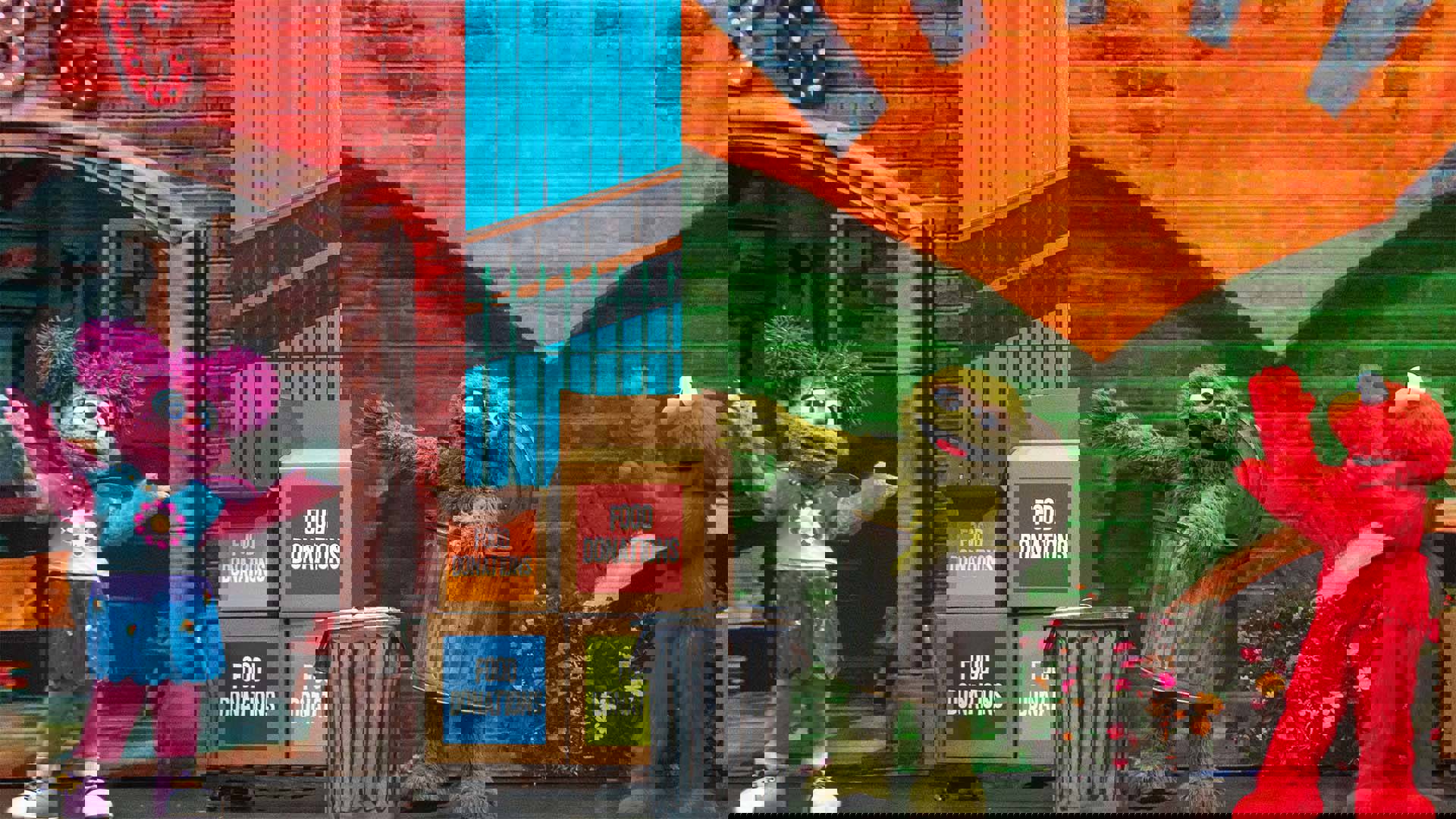 Sesame Street characters Abby Cadabby, Oscar the Grouch and Elmo perform on stage with colorful backdrops and boxes labeled “Food Donations.”
